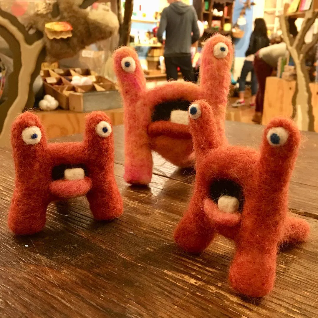 Three orange-red H-shaped felted monsters with eyes on stalks and open mouths, on a wooden table