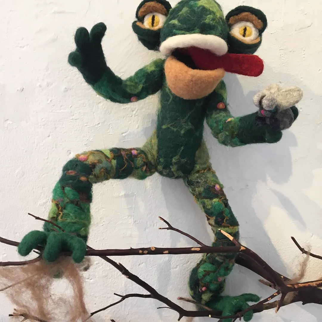 A large green felted frog creature with a red tongue, climbing a branch on a white wall and holding a felted fly