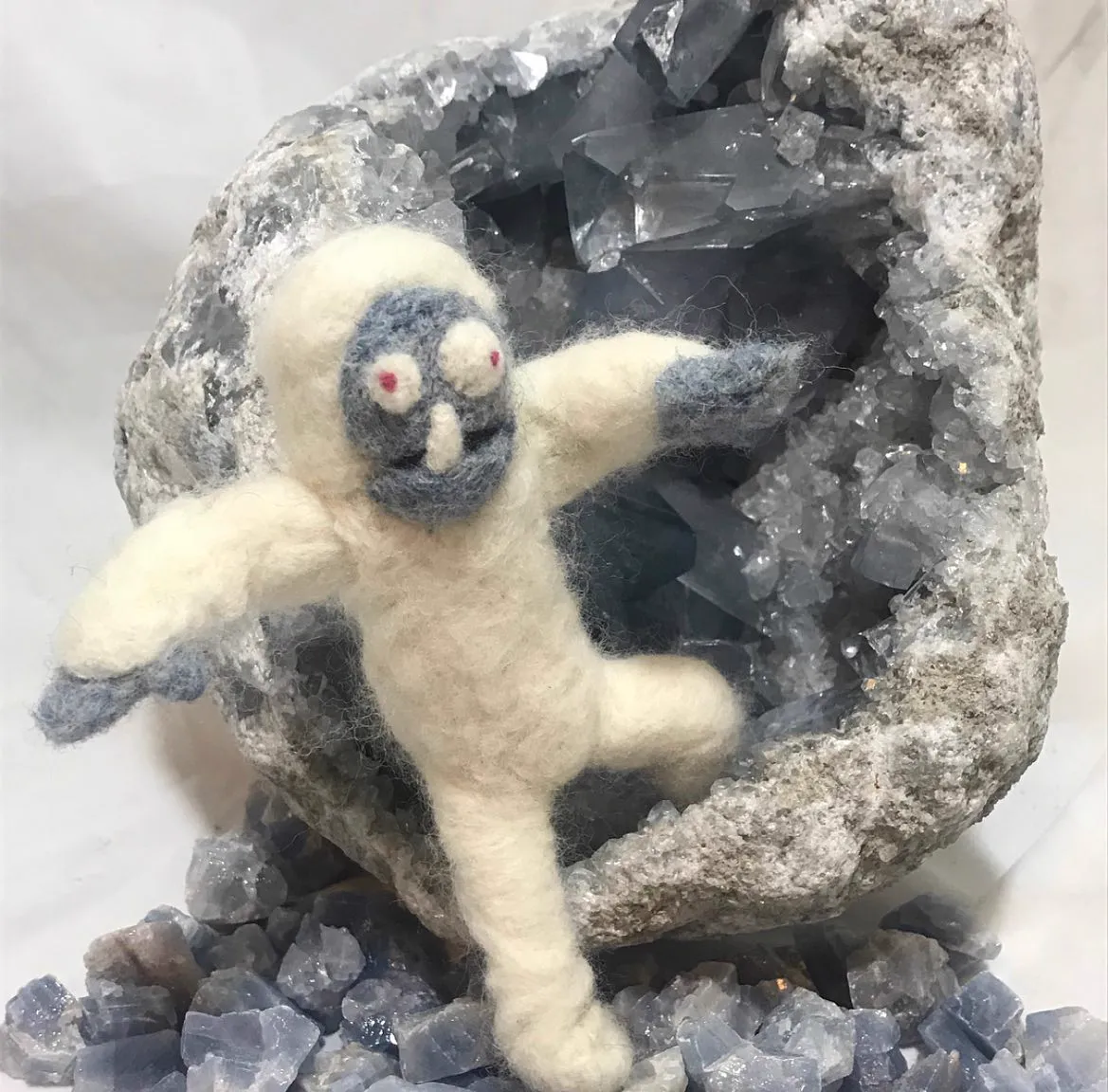 Small white felted yeti posed mid-leap in front of a large blue celestite geode
