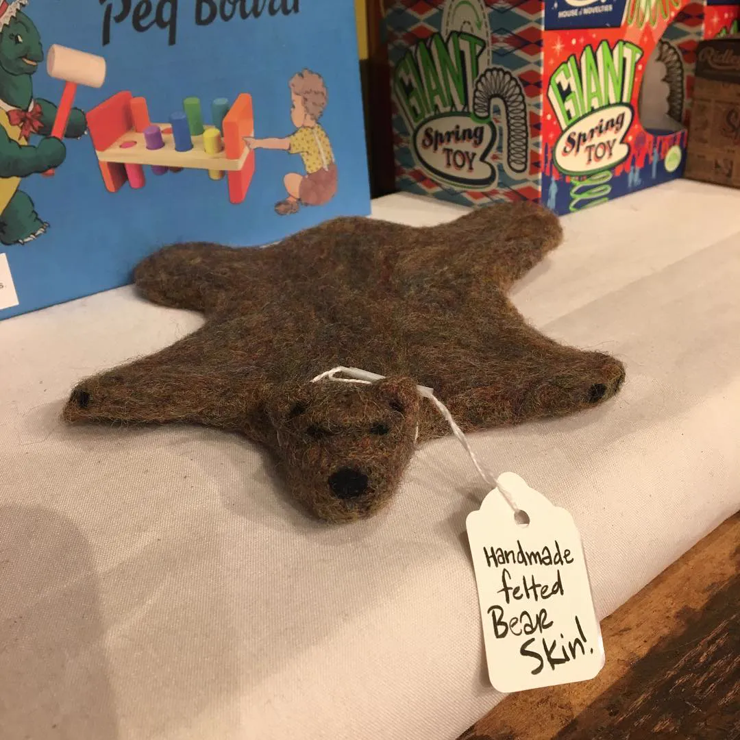A tiny felted brown bear-skin rug on a shelf with a handwritten tag: Handmade felted bear skin!