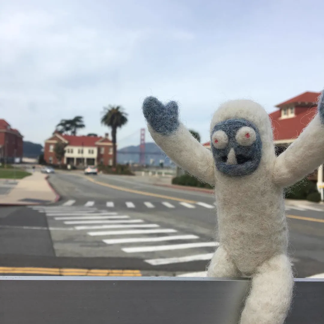 Freddi Yeti waving on a railing in the Presidio with the Golden Gate Bridge behind