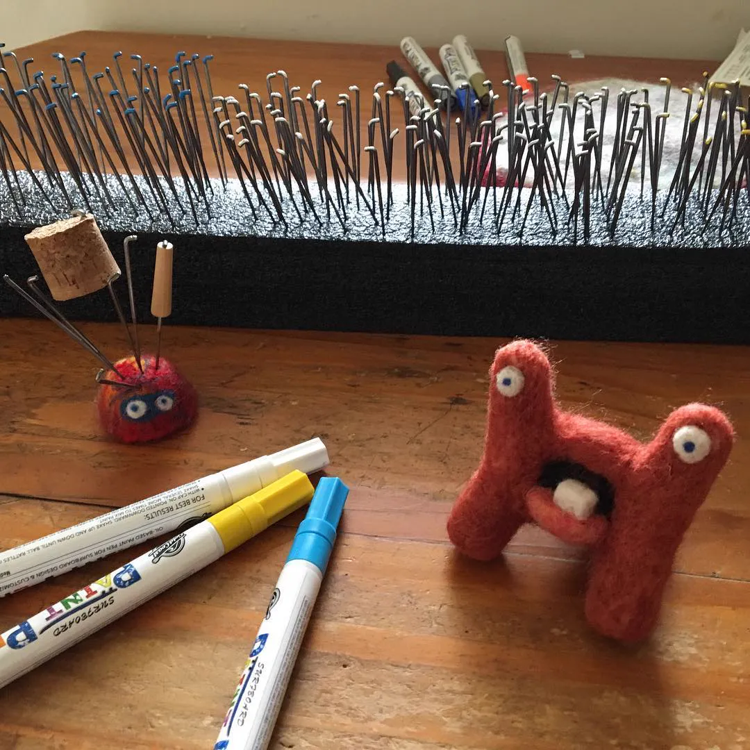 Rows of felting needles standing in a foam block, their tips painted different colors, with paint pens and a finished Hectrix on the table